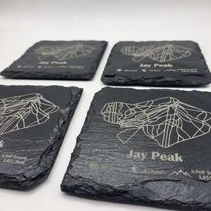 May include: Four black slate coasters with a white outline of Jay Peak, a mountain in Vermont, and the text "Jay Peak" and "3,968 Summit 1,851 Base".