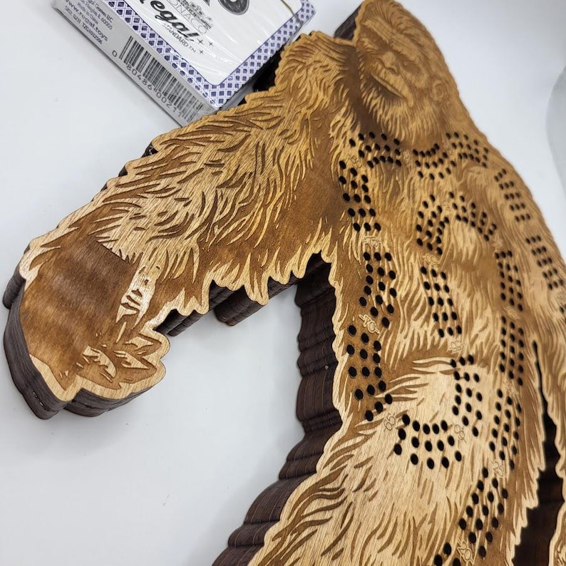 May include: A wooden Bigfoot-shaped game board with a brown stain. The board has a series of holes for playing a game. The board is made of layered wood.