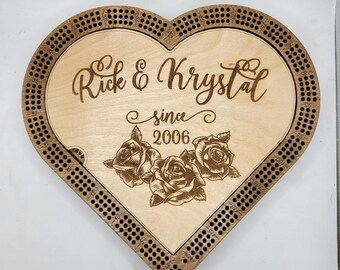 May include: Heart-shaped wooden cribbage board with the names "Rick & Krystal" and the year "2006" engraved. Features a floral design of three roses. The board has a border with holes for pegs.