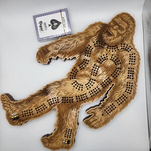 May include: A wooden Bigfoot game board with a brown stain. The board is shaped like a Bigfoot with a detailed design. The board has a series of holes for playing the game.