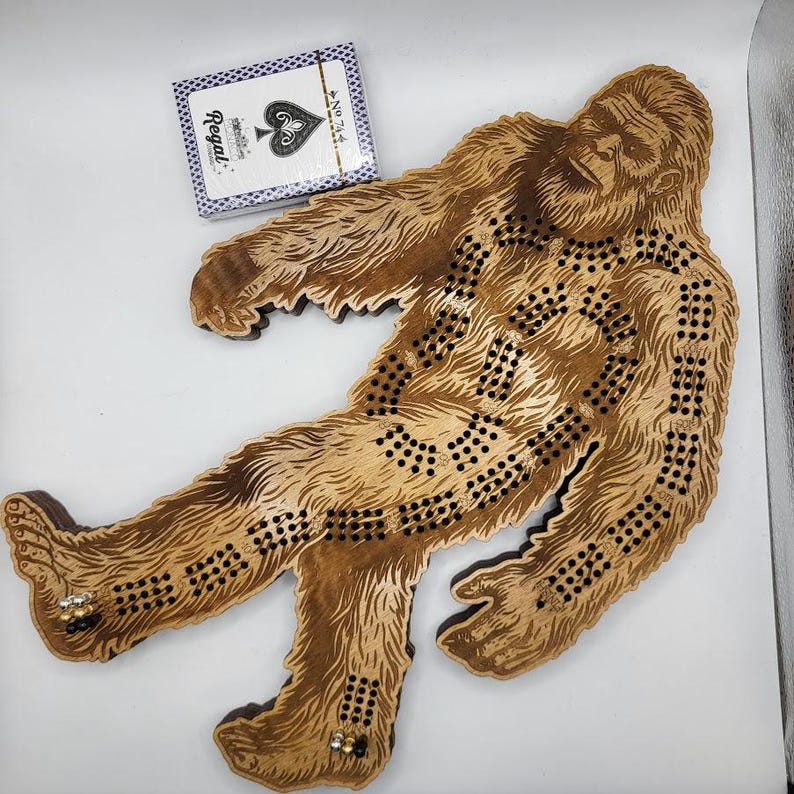 May include: A wooden Bigfoot game board with a brown stain. The board is shaped like a Bigfoot with a detailed design. The board has a series of holes for playing the game.