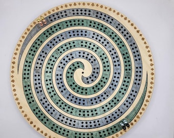 May include: A circular wooden cribbage board featuring a spiral track with alternating green and blue stained sections. The board includes numbered scoring holes, card suit symbols along the outer edge, and two sets of metal pegs at the start positions.
