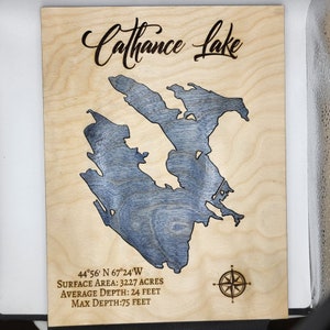 May include: A wooden sign with a laser-cut map of Cathance Lake, Maine. The sign includes the lake's coordinates, surface area, average depth, and maximum depth. The sign is a light brown color with a compass rose in the bottom right corner.