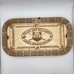 May include: A wooden game board with a pool table design. The board is engraved with the text "Bears Den Billiards Trapt Escape Rooms".