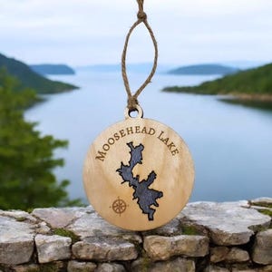 May include: A wooden ornament with a cutout of Moosehead Lake and the text "Moosehead Lake" around the edge. The ornament is hanging from a brown twine string.