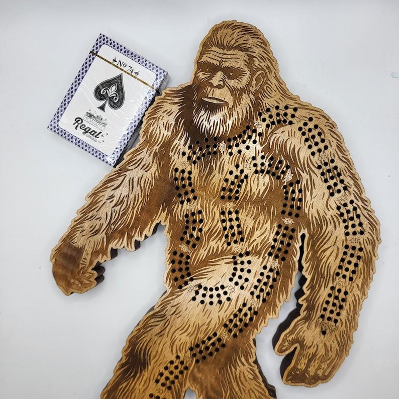 May include: Wooden Bigfoot peg game with a brown stain finish. The game board is shaped like a Bigfoot standing with its arms outstretched. The game includes a playing card with the number 74 and the suit of spades.