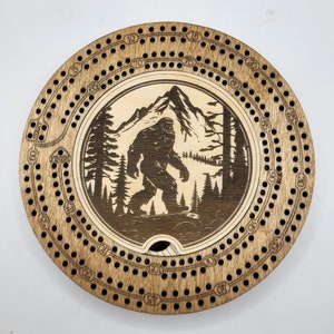 May include: A wooden cribbage board with a circular design. The board has a center circle with a silhouette of Bigfoot walking through a forest with a mountain in the background. The outer circle has numbers from 1 to 120.