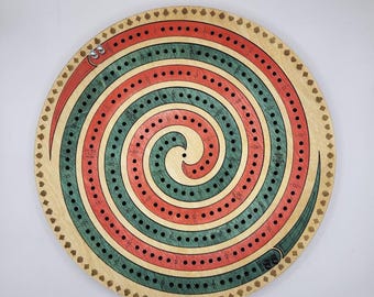 May include: A circular wooden cribbage board featuring a spiral track design with alternating red and teal stained sections. The board includes drilled holes for scoring, numbered markings, and two metal pegs positioned at the start of the track.