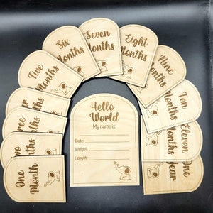 May include: A collection of wooden baby milestone cards. The cards are light brown with engraved text and an elephant graphic. The set includes cards for months one to twelve, plus a "Hello World" card for baby's details.