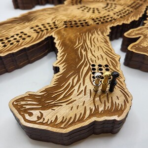 May include: A wooden game board shaped like a Bigfoot with a brown stain finish. The board has a grid of holes and is made of multiple layers of wood. There are three pegs in the holes, one gold, one silver, and one black.
