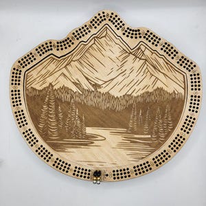 May include: A wooden cribbage board featuring a mountain and lake scene. The board has a brown stain and a black border with numbered holes. The board is shaped like a mountain range.