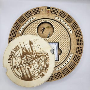 May include: A circular wooden cribbage board with a removable lid featuring a laser-engraved lighthouse, compass, and the text "ALWAYS TAKE THE SCENIC ROUTE." The board includes numbered scoring tracks, metal pegs in various colours, and a storage compartment.
