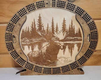 May include: A wooden cribbage board with a laser-engraved image of a moose standing in a lake surrounded by trees. The board has a circular shape with a raised edge and a flat playing surface. The board is made of dark wood with a natural finish.