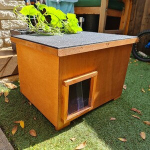 May include: A wooden pet house with a brown stain finish and a clear plastic door. The house has a flat roof and a small opening for the pet to enter.