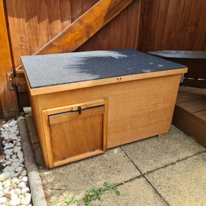 May include: A wooden cat house with a black, asphalt shingle roof. The rectangular house has a small wooden door with a latch. The cat house is made of light-colored wood and sits on a concrete patio. The dimensions are approximately 75 cm long, 50 cm wide, and 50 cm high.