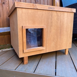 May include: A wooden cat house with a DINGO logo on the top. The house has a small window and is sitting on a wooden deck.