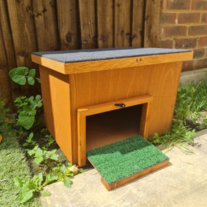 May include: A wooden hedgehog house with a green artificial grass ramp. The house has a black roof and a small door.