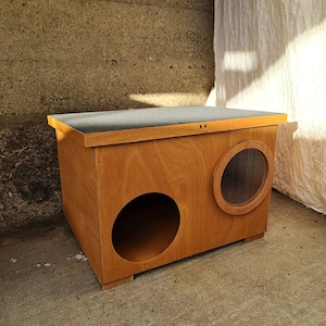 May include: A wooden pet house with a flat, gray roof and two circular openings. One opening is a large hole, and the other is a smaller, circular window. The house is made of light-colored wood and sits on a concrete surface.