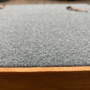 May include: A close-up of a gray, textured surface with a wooden frame. The surface is smooth and appears to be made of felt.