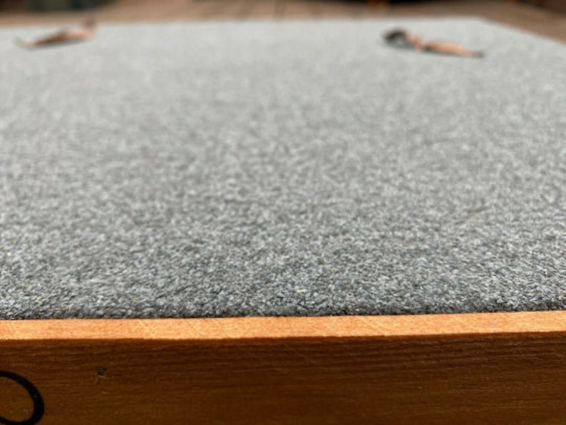 May include: A close-up of a gray, textured surface with a wooden frame. The surface is smooth and appears to be made of felt.