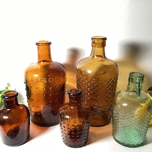 May include: A collection of five glass bottles including three amber bottles with embossed LYSOL text and dot patterns, one tall amber bottle with a diamond texture, and one clear green bottle with a diamond pattern, all with narrow necks.