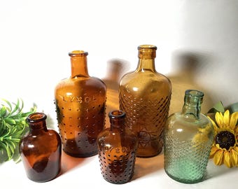 Puede incluir: Una colección de cinco botellas de vidrio que incluye tres botellas ámbar con texto LYSOL en relieve y patrones de puntos, una botella ámbar alta con textura de diamante y una botella verde claro con patrón de diamante, todas con cuello estrecho.