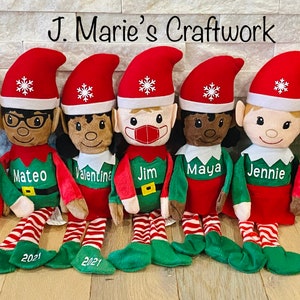May include: Eight personalized elf dolls with red and green outfits, each with a different name and the year 2021 embroidered on their legs. The elves are sitting on a wooden surface.