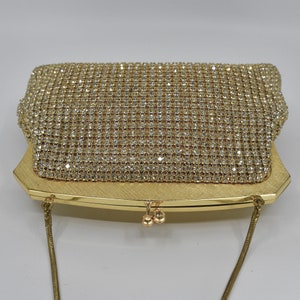 May include: A gold-toned evening clutch with a sparkling rhinestone mesh overlay. The rectangular bag features a brushed gold frame and a delicate chain strap. The clasp has two small gold spheres. A glamorous accessory.