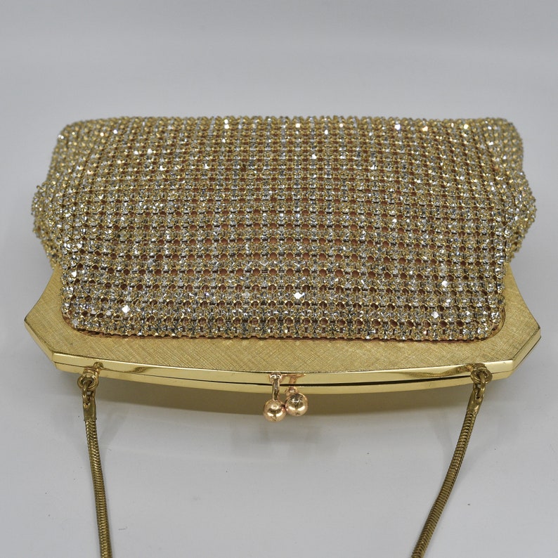 May include: A gold-toned evening clutch with a sparkling rhinestone mesh overlay. The rectangular bag features a brushed gold frame and a delicate chain strap. The clasp has two small gold spheres. A glamorous accessory.