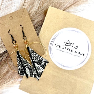 May include: Dangle earrings featuring black hooks, gold-tone hoops, and triangular pendants with a black and white snakeskin pattern. The earrings are presented on a brown card, alongside a sticker with the text "THE STYLE MODE".