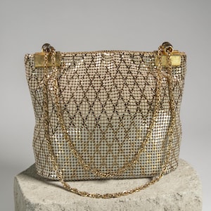 May include: A vintage gold mesh handbag with a diamond pattern. The bag has a gold chain strap and gold hardware. The bag is on a stone surface. The mesh material gives the bag a shimmering appearance.