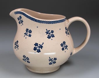 May include: Cream ceramic jug with a rounded body and a handle. The jug is decorated with blue floral patterns and a blue rope design around the rim. It has a spout for easy pouring.