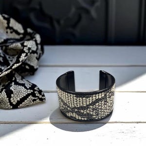 May include: A wide cuff bracelet with a snakeskin pattern in black and white. The bracelet has a black interior and trim. A scarf with a similar pattern is visible in the background.