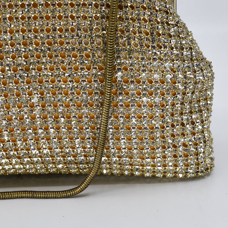 May include: A close-up of a vintage, gold-toned evening bag. The bag is covered in sparkling rhinestones, creating a shimmering effect. A gold-colored chain strap is visible. The interior appears to be a warm, golden color.