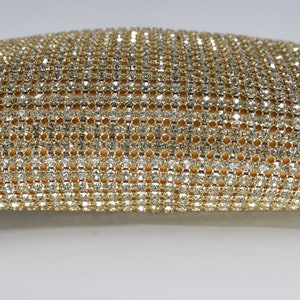May include: Close-up of a gold and silver rhinestone mesh fabric. The mesh is densely packed with small, sparkling rhinestones, creating a shimmering effect. The fabric has a warm, golden-yellow backing visible through the mesh.