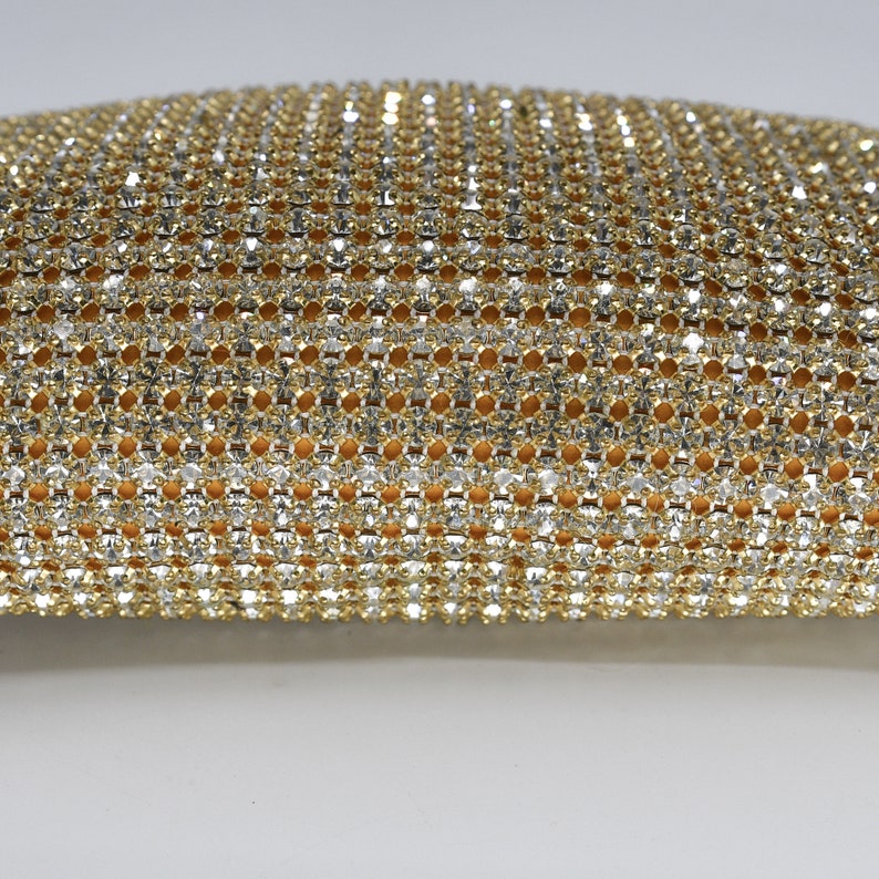 May include: Close-up of a gold and silver rhinestone mesh fabric. The mesh is densely packed with small, sparkling rhinestones, creating a shimmering effect. The fabric has a warm, golden-yellow backing visible through the mesh.
