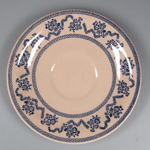 May include: A beige ceramic plate featuring a blue floral and ribbon pattern around the rim. The plate has a rope-style border and a smooth, slightly recessed centre. The floral design is repeated around the plate's edge.