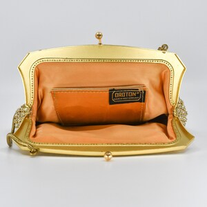 May include: Open, gold-toned vintage handbag with a peach-colored interior. The frame is embellished with small, clear rhinestones. The label inside reads "OROTON MADE IN WEST GERMANY". The bag has a small chain handle and a clasp closure.