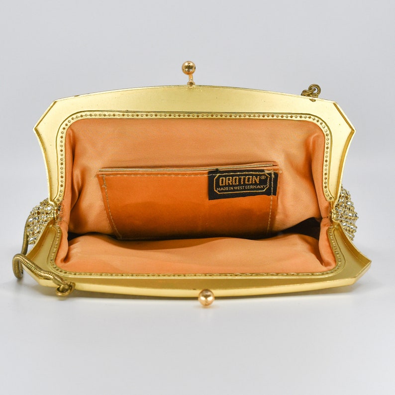 May include: Open, gold-toned vintage handbag with a peach-colored interior. The frame is embellished with small, clear rhinestones. The label inside reads "OROTON MADE IN WEST GERMANY". The bag has a small chain handle and a clasp closure.