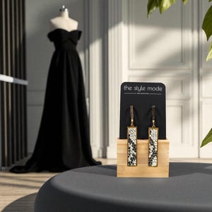 May include: Rectangular earrings with a black and white pattern, displayed on a wooden stand. The earrings have gold-coloured frames. A black dress on a mannequin is in the background. The text "the style mode" is visible on the display card.