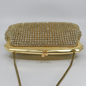 May include: A gold-toned evening clutch with a rectangular shape. The top flap is covered in sparkling rhinestones, while the body has a textured finish. A delicate gold chain strap is attached. The clasp features two small, round gold accents.