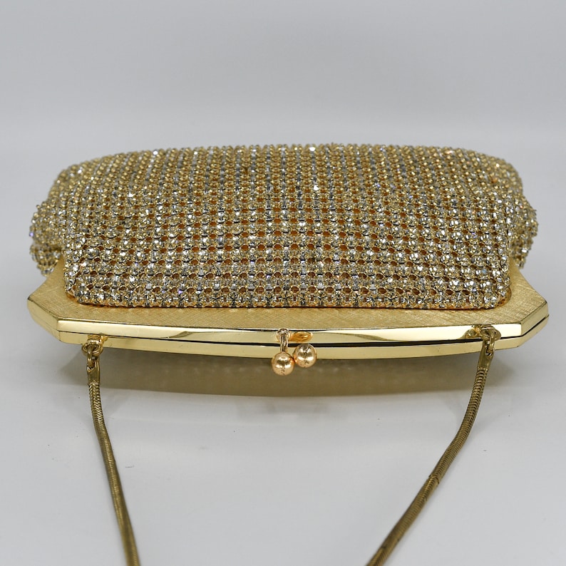 May include: A gold-toned evening clutch with a rectangular shape. The top flap is covered in sparkling rhinestones, while the body has a textured finish. A delicate gold chain strap is attached. The clasp features two small, round gold accents.