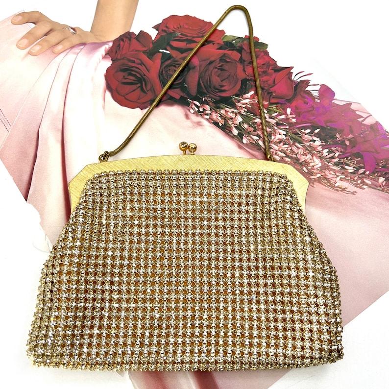 May include: A vintage gold-toned evening bag with a delicate chain handle. The bag is covered in a shimmering, textured pattern. The clasp is a small, decorative gold tone. The background includes a bouquet of red roses.
