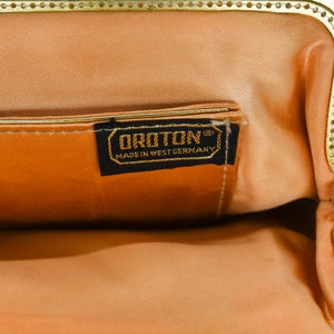 May include: Open view of a vintage peach-colored handbag with a gold-toned clasp. The interior features a matching peach lining and a small pocket. A black label with gold lettering reads "OROTON MADE IN WEST GERMANY."
