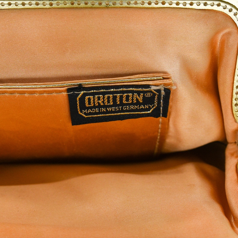May include: Open view of a vintage peach-colored handbag with a gold-toned clasp. The interior features a matching peach lining and a small pocket. A black label with gold lettering reads "OROTON MADE IN WEST GERMANY."