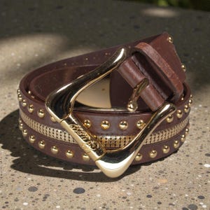 May include: A brown leather belt with gold-coloured metal accents. The belt has a large gold buckle and is decorated with small gold studs and a gold mesh strip. The belt is coiled on a speckled surface.