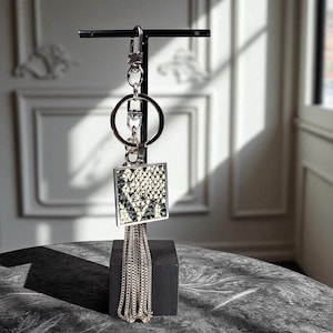 May include: A silver keychain with a square pendant featuring a black and white snakeskin pattern. The pendant is framed in silver and has a fringe of silver chains. The keychain is attached to a silver ring and clasp.