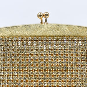 May include: Close-up of a vintage gold-toned evening bag. The bag features a sparkling, rhinestone-studded mesh body and a textured gold frame. The clasp is a pair of small, round gold spheres. The bag is set against a white background.