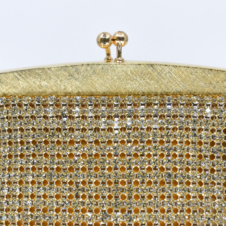 May include: Close-up of a vintage gold-toned evening bag. The bag features a sparkling, rhinestone-studded mesh body and a textured gold frame. The clasp is a pair of small, round gold spheres. The bag is set against a white background.