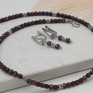 Könnte beinhalten: Ein Schmuckset bestehend aus einer Halskette und einem Paar Ohrringen. Die Kette besteht aus kleinen, facettierten dunkelroten Perlen, die mit metallisch silberfarbenen Zwischenelementen abwechseln. Die passenden Ohrringe haben eine silberfarbene Metallfassung.